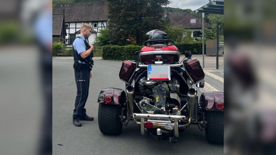 Ein Polizeibeamter steht auf einer Straße neben einem dreirädrigen Motorrad mit breiter Hinterachse. Das Fahrzeug ist von hinten zu sehen und mit Seitenkoffern sowie einem Topcase ausgestattet. Im Hintergrund befinden sich Wohnhäuser und eine Bushaltestelle.