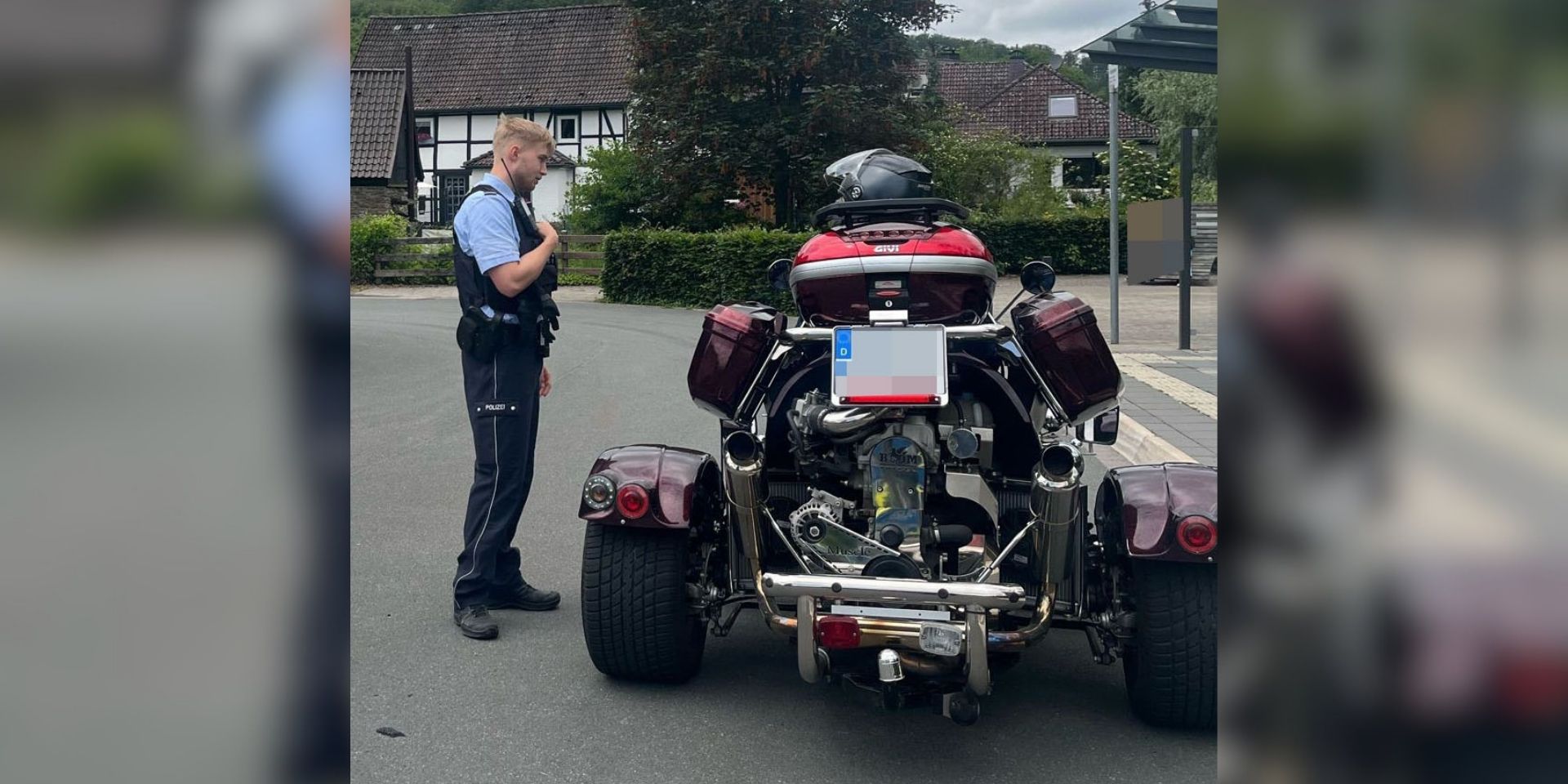 Ein Polizeibeamter steht auf einer Straße neben einem dreirädrigen Motorrad mit breiter Hinterachse. Das Fahrzeug ist von hinten zu sehen und mit Seitenkoffern sowie einem Topcase ausgestattet. Im Hintergrund befinden sich Wohnhäuser und eine Bushaltestelle.