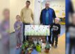 Zwei Männer und zwei Kinder stehen in den Räumen einer Kindertageseinrichtung hinter einer dekorierten Fotowand. Vor ihnen sind kleine Blumentöpfe, eine grüne Gießkanne, Metalleimer und Dankeskarten arrangiert. Die Fotowand zeigt eine bunte Blumenwiese mit Schmetterlingen. Das Bild entstand anlässlich des Tags der Kinderbetreuung und zeigt eine symbolische Dankesaktion für die Mitarbeitenden der Einrichtung.