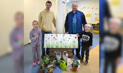 Zwei Männer und zwei Kinder stehen in den Räumen einer Kindertageseinrichtung hinter einer dekorierten Fotowand. Vor ihnen sind kleine Blumentöpfe, eine grüne Gießkanne, Metalleimer und Dankeskarten arrangiert. Die Fotowand zeigt eine bunte Blumenwiese mit Schmetterlingen. Das Bild entstand anlässlich des Tags der Kinderbetreuung und zeigt eine symbolische Dankesaktion für die Mitarbeitenden der Einrichtung.