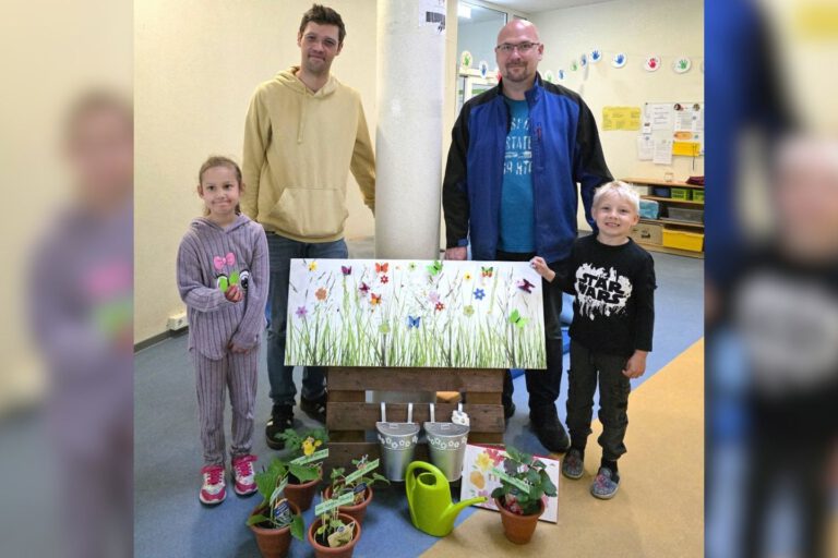Zwei Männer und zwei Kinder stehen in den Räumen einer Kindertageseinrichtung hinter einer dekorierten Fotowand. Vor ihnen sind kleine Blumentöpfe, eine grüne Gießkanne, Metalleimer und Dankeskarten arrangiert. Die Fotowand zeigt eine bunte Blumenwiese mit Schmetterlingen. Das Bild entstand anlässlich des Tags der Kinderbetreuung und zeigt eine symbolische Dankesaktion für die Mitarbeitenden der Einrichtung.