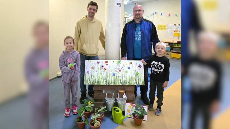 Zwei Männer und zwei Kinder stehen in den Räumen einer Kindertageseinrichtung hinter einer dekorierten Fotowand. Vor ihnen sind kleine Blumentöpfe, eine grüne Gießkanne, Metalleimer und Dankeskarten arrangiert. Die Fotowand zeigt eine bunte Blumenwiese mit Schmetterlingen. Das Bild entstand anlässlich des Tags der Kinderbetreuung und zeigt eine symbolische Dankesaktion für die Mitarbeitenden der Einrichtung.