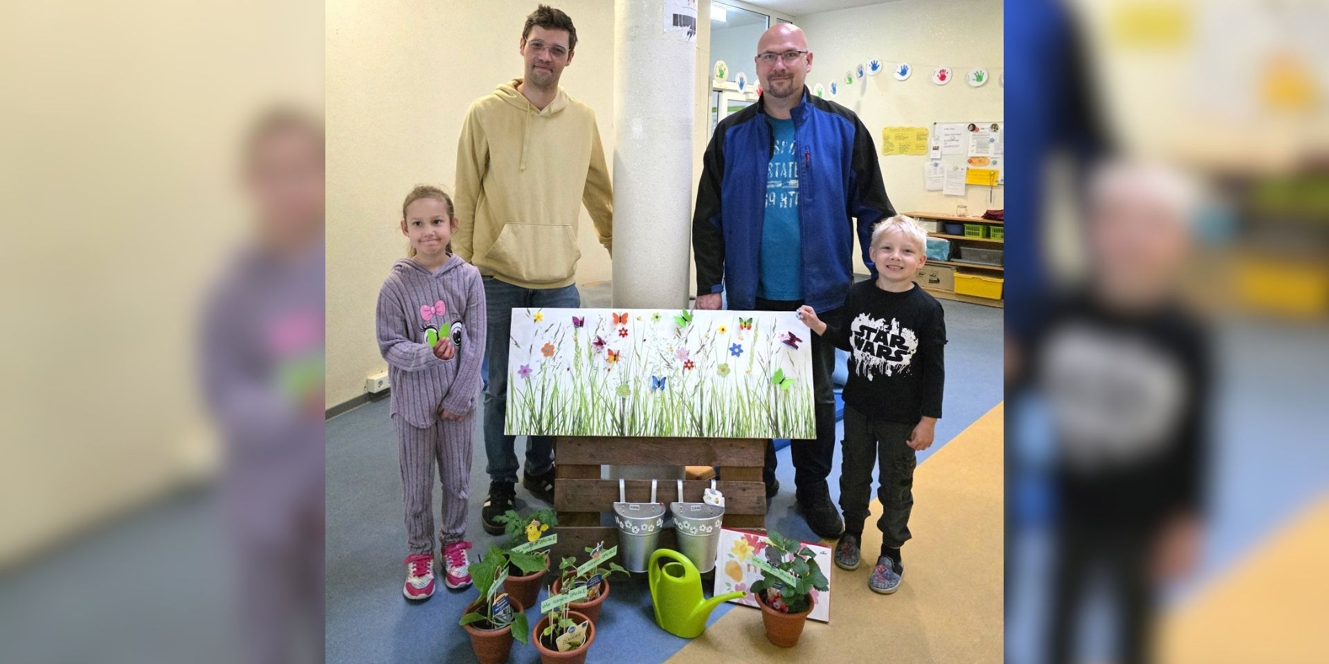 Zwei Männer und zwei Kinder stehen in den Räumen einer Kindertageseinrichtung hinter einer dekorierten Fotowand. Vor ihnen sind kleine Blumentöpfe, eine grüne Gießkanne, Metalleimer und Dankeskarten arrangiert. Die Fotowand zeigt eine bunte Blumenwiese mit Schmetterlingen. Das Bild entstand anlässlich des Tags der Kinderbetreuung und zeigt eine symbolische Dankesaktion für die Mitarbeitenden der Einrichtung.