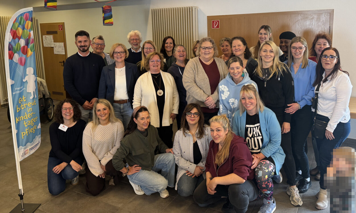 Gruppenfoto von rund 25 Teilnehmenden einer Veranstaltung zur Kindertagespflege in einem Gemeinschaftsraum. Die Gruppe steht und kniet für das Foto, daneben ist ein Banner mit der Aufschrift „Gut betreut in Kindertagespflege“ zu sehen. Im Vordergrund rechts ist ein Kind aus Datenschutzgründen verpixelt.