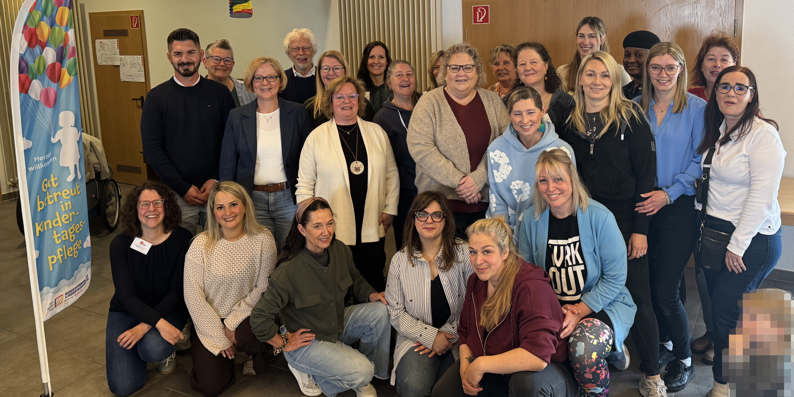 Gruppenfoto von rund 25 Teilnehmenden einer Veranstaltung zur Kindertagespflege in einem Gemeinschaftsraum. Die Gruppe steht und kniet für das Foto, daneben ist ein Banner mit der Aufschrift „Gut betreut in Kindertagespflege“ zu sehen. Im Vordergrund rechts ist ein Kind aus Datenschutzgründen verpixelt.