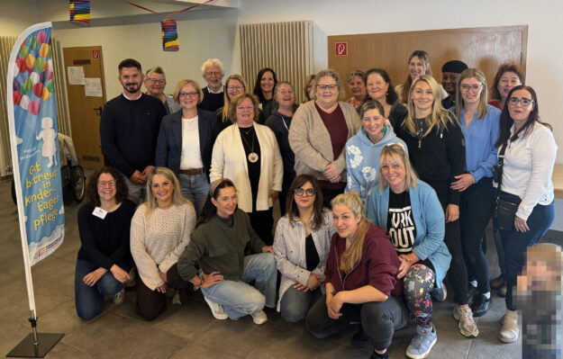 Gruppenfoto von rund 25 Teilnehmenden einer Veranstaltung zur Kindertagespflege in einem Gemeinschaftsraum. Die Gruppe steht und kniet für das Foto, daneben ist ein Banner mit der Aufschrift „Gut betreut in Kindertagespflege“ zu sehen. Im Vordergrund rechts ist ein Kind aus Datenschutzgründen verpixelt.