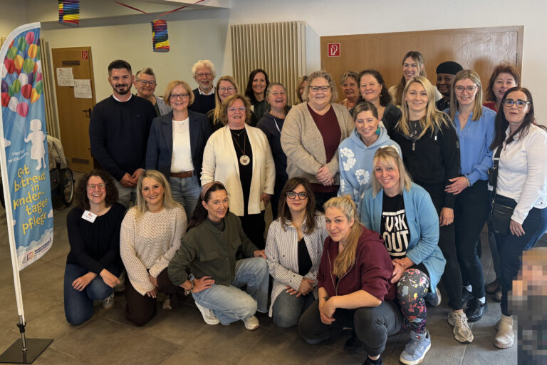 Gruppenfoto von rund 25 Teilnehmenden einer Veranstaltung zur Kindertagespflege in einem Gemeinschaftsraum. Die Gruppe steht und kniet für das Foto, daneben ist ein Banner mit der Aufschrift „Gut betreut in Kindertagespflege“ zu sehen. Im Vordergrund rechts ist ein Kind aus Datenschutzgründen verpixelt.