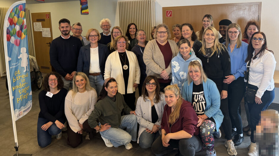 Gruppenfoto von rund 25 Teilnehmenden einer Veranstaltung zur Kindertagespflege in einem Gemeinschaftsraum. Die Gruppe steht und kniet für das Foto, daneben ist ein Banner mit der Aufschrift „Gut betreut in Kindertagespflege“ zu sehen. Im Vordergrund rechts ist ein Kind aus Datenschutzgründen verpixelt.