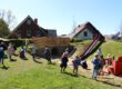 Mehrere Kinder spielen auf einem großzügigen Außengelände mit Rutsche, Hügel und Spielgeräten. Im Hintergrund stehen Einfamilienhäuser, während die Kinder über die Wiese laufen, klettern und gemeinsam den Spielplatz nutzen. Die Aufnahme vermittelt eine lebendige und fröhliche Atmosphäre.