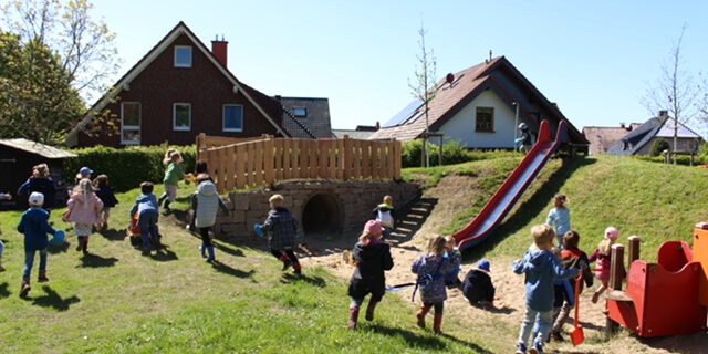 Mehrere Kinder spielen auf einem großzügigen Außengelände mit Rutsche, Hügel und Spielgeräten. Im Hintergrund stehen Einfamilienhäuser, während die Kinder über die Wiese laufen, klettern und gemeinsam den Spielplatz nutzen. Die Aufnahme vermittelt eine lebendige und fröhliche Atmosphäre.
