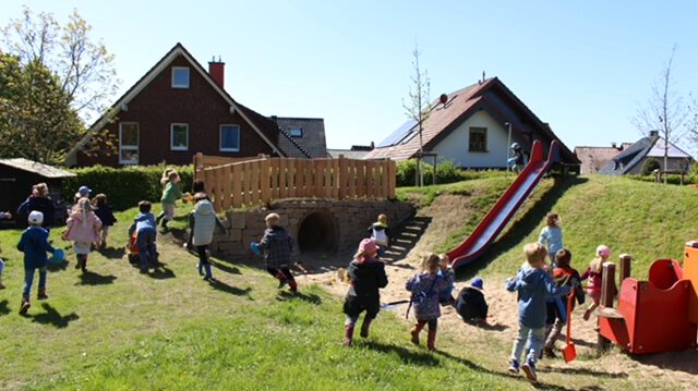 Mehrere Kinder spielen auf einem großzügigen Außengelände mit Rutsche, Hügel und Spielgeräten. Im Hintergrund stehen Einfamilienhäuser, während die Kinder über die Wiese laufen, klettern und gemeinsam den Spielplatz nutzen. Die Aufnahme vermittelt eine lebendige und fröhliche Atmosphäre.