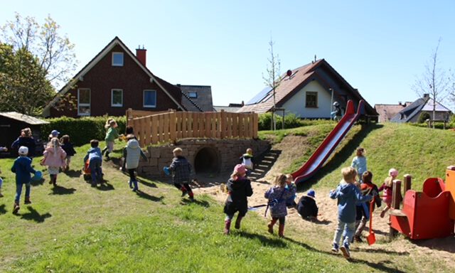 Mehrere Kinder spielen auf einem großzügigen Außengelände mit Rutsche, Hügel und Spielgeräten. Im Hintergrund stehen Einfamilienhäuser, während die Kinder über die Wiese laufen, klettern und gemeinsam den Spielplatz nutzen. Die Aufnahme vermittelt eine lebendige und fröhliche Atmosphäre.