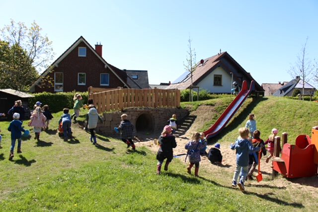Mehrere Kinder spielen auf einem großzügigen Außengelände mit Rutsche, Hügel und Spielgeräten. Im Hintergrund stehen Einfamilienhäuser, während die Kinder über die Wiese laufen, klettern und gemeinsam den Spielplatz nutzen. Die Aufnahme vermittelt eine lebendige und fröhliche Atmosphäre.
