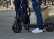 Ein E-Scooter-Fahrer steht mit seinem Roller im Gespräch mit einem Polizeibeamten auf einem Platz im Freien. Der schwarze E-Scooter ist gut sichtbar, am Lenker hängt ein Rucksack. Im Hintergrund stehen Tische und Stühle einer Außengastronomie. Das Bild zeigt eine Polizeikontrolle im öffentlichen Raum.