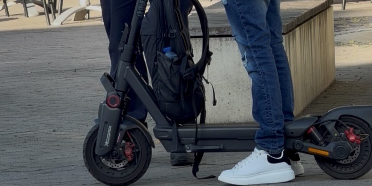 Ein E-Scooter-Fahrer steht mit seinem Roller im Gespräch mit einem Polizeibeamten auf einem Platz im Freien. Der schwarze E-Scooter ist gut sichtbar, am Lenker hängt ein Rucksack. Im Hintergrund stehen Tische und Stühle einer Außengastronomie. Das Bild zeigt eine Polizeikontrolle im öffentlichen Raum.
