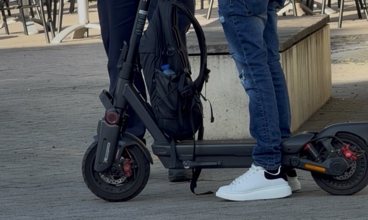 Ein E-Scooter-Fahrer steht mit seinem Roller im Gespräch mit einem Polizeibeamten auf einem Platz im Freien. Der schwarze E-Scooter ist gut sichtbar, am Lenker hängt ein Rucksack. Im Hintergrund stehen Tische und Stühle einer Außengastronomie. Das Bild zeigt eine Polizeikontrolle im öffentlichen Raum.