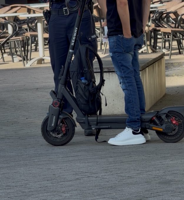 Ein E-Scooter-Fahrer steht mit seinem Roller im Gespräch mit einem Polizeibeamten auf einem Platz im Freien. Der schwarze E-Scooter ist gut sichtbar, am Lenker hängt ein Rucksack. Im Hintergrund stehen Tische und Stühle einer Außengastronomie. Das Bild zeigt eine Polizeikontrolle im öffentlichen Raum.