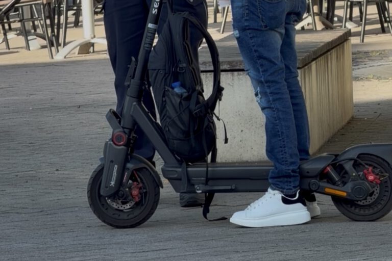 Ein E-Scooter-Fahrer steht mit seinem Roller im Gespräch mit einem Polizeibeamten auf einem Platz im Freien. Der schwarze E-Scooter ist gut sichtbar, am Lenker hängt ein Rucksack. Im Hintergrund stehen Tische und Stühle einer Außengastronomie. Das Bild zeigt eine Polizeikontrolle im öffentlichen Raum.