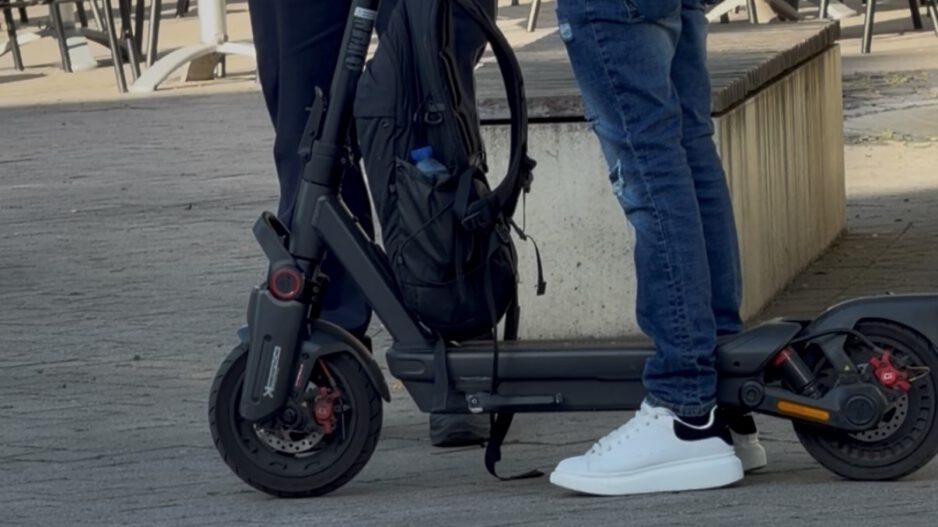 Ein E-Scooter-Fahrer steht mit seinem Roller im Gespräch mit einem Polizeibeamten auf einem Platz im Freien. Der schwarze E-Scooter ist gut sichtbar, am Lenker hängt ein Rucksack. Im Hintergrund stehen Tische und Stühle einer Außengastronomie. Das Bild zeigt eine Polizeikontrolle im öffentlichen Raum.