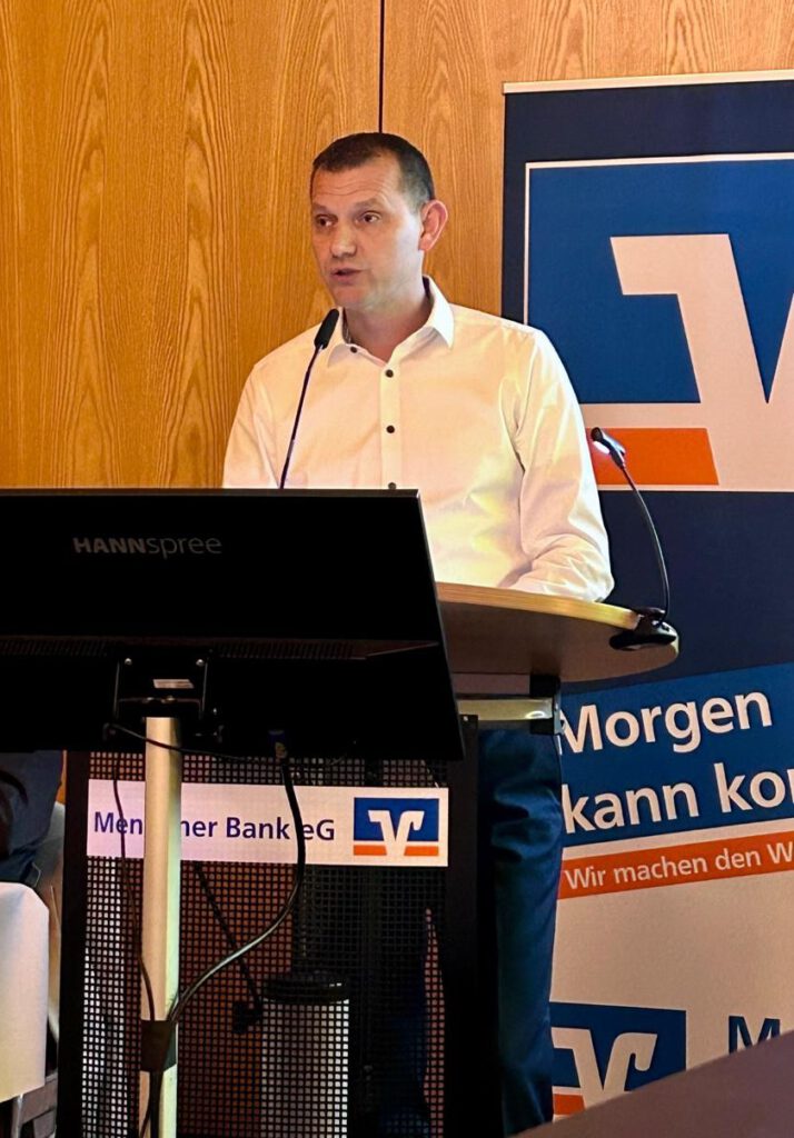 Ein Mann in weißem Hemd steht an einem Rednerpult und hält einen Vortrag. Vor ihm befinden sich ein Bildschirm und Mikrofone, im Hintergrund ist ein Banner der Mendener Bank zu sehen.