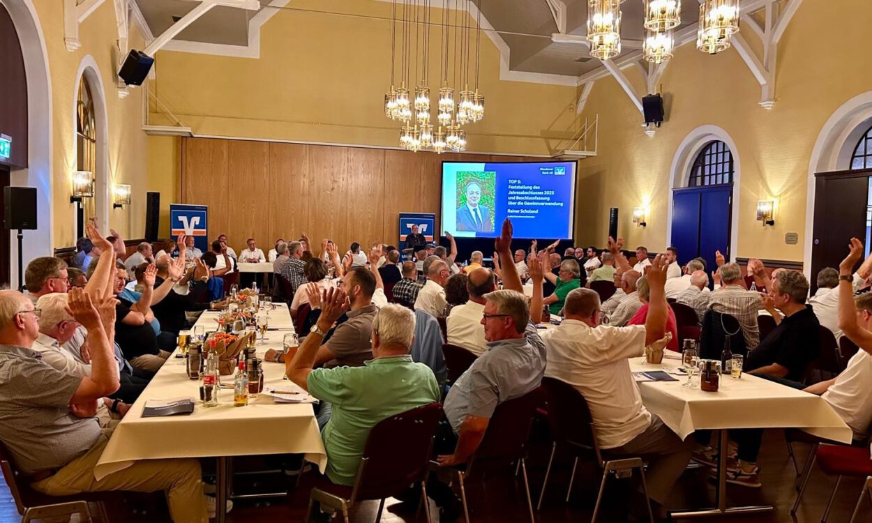 In einem festlich beleuchteten Veranstaltungssaal sitzen zahlreiche Mitglieder einer Genossenschaft an langen Tischen. Während einer Abstimmung heben viele der Anwesenden die Hand. Im Hintergrund ist eine Präsentationsfolie mit Informationen zum Jahresabschluss und zur Beschlussfassung zu sehen. Die Aufnahme zeigt eine Mitgliederversammlung mit reger Beteiligung.