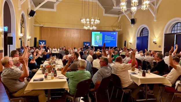 In einem festlich beleuchteten Veranstaltungssaal sitzen zahlreiche Mitglieder einer Genossenschaft an langen Tischen. Während einer Abstimmung heben viele der Anwesenden die Hand. Im Hintergrund ist eine Präsentationsfolie mit Informationen zum Jahresabschluss und zur Beschlussfassung zu sehen. Die Aufnahme zeigt eine Mitgliederversammlung mit reger Beteiligung.