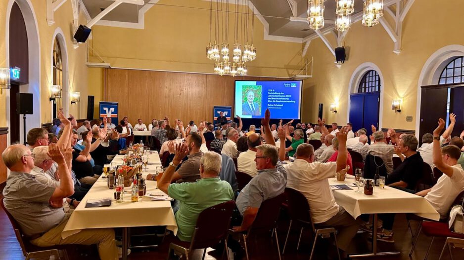 In einem festlich beleuchteten Veranstaltungssaal sitzen zahlreiche Mitglieder einer Genossenschaft an langen Tischen. Während einer Abstimmung heben viele der Anwesenden die Hand. Im Hintergrund ist eine Präsentationsfolie mit Informationen zum Jahresabschluss und zur Beschlussfassung zu sehen. Die Aufnahme zeigt eine Mitgliederversammlung mit reger Beteiligung.