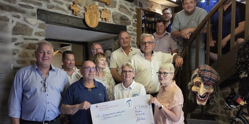 Eine Gruppe von Frauen und Männern steht in einem historischen Fachwerkgebäude für ein Gruppenfoto zusammen. Im Vordergrund halten mehrere Personen einen symbolischen Spendenscheck in die Kamera. Im Hintergrund führen eine Holztreppe und Natursteinwände durch den Raum, daneben steht eine große Hexenfigur als Dekoration. Die Aufnahme zeigt die Übergabe einer Spende im Beisein der beteiligten Vereinsmitglieder und Unterstützer.