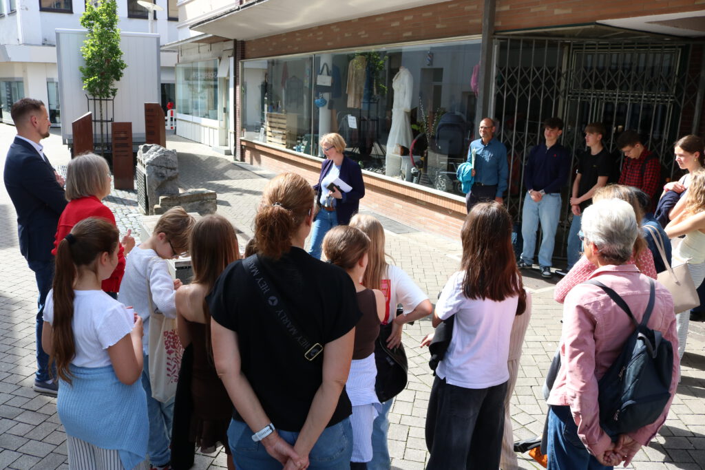 Eine Gruppe von Jugendlichen und Erwachsenen steht an einem sonnigen Tag in einer Fußgängerzone und hört einer Frau zu, die vor ihnen spricht. Die Teilnehmer haben sich halbkreisförmig um mehrere Gedenksteine und Informationsstelen versammelt. Im Hintergrund befinden sich Schaufenster eines Geschäfts. Die Szene wirkt wie eine historische Stadtführung oder eine Gedenkveranstaltung an einem Erinnerungsort im öffentlichen Raum