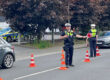 Zwei Polizeibeamte führen auf einer Straße eine Verkehrskontrolle durch. Zwischen mehreren orange-weißen Leitkegeln regeln sie den Verkehr und weisen Fahrzeuge ein. Im Hintergrund sind ein Streifenwagen sowie weitere Autos zu sehen. Die Aufnahme zeigt eine polizeiliche Kontrollstelle im öffentlichen Straßenverkehr.