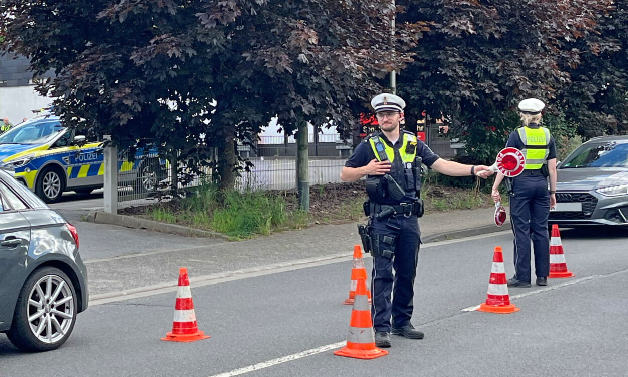 Zwei Polizeibeamte führen auf einer Straße eine Verkehrskontrolle durch. Zwischen mehreren orange-weißen Leitkegeln regeln sie den Verkehr und weisen Fahrzeuge ein. Im Hintergrund sind ein Streifenwagen sowie weitere Autos zu sehen. Die Aufnahme zeigt eine polizeiliche Kontrollstelle im öffentlichen Straßenverkehr.