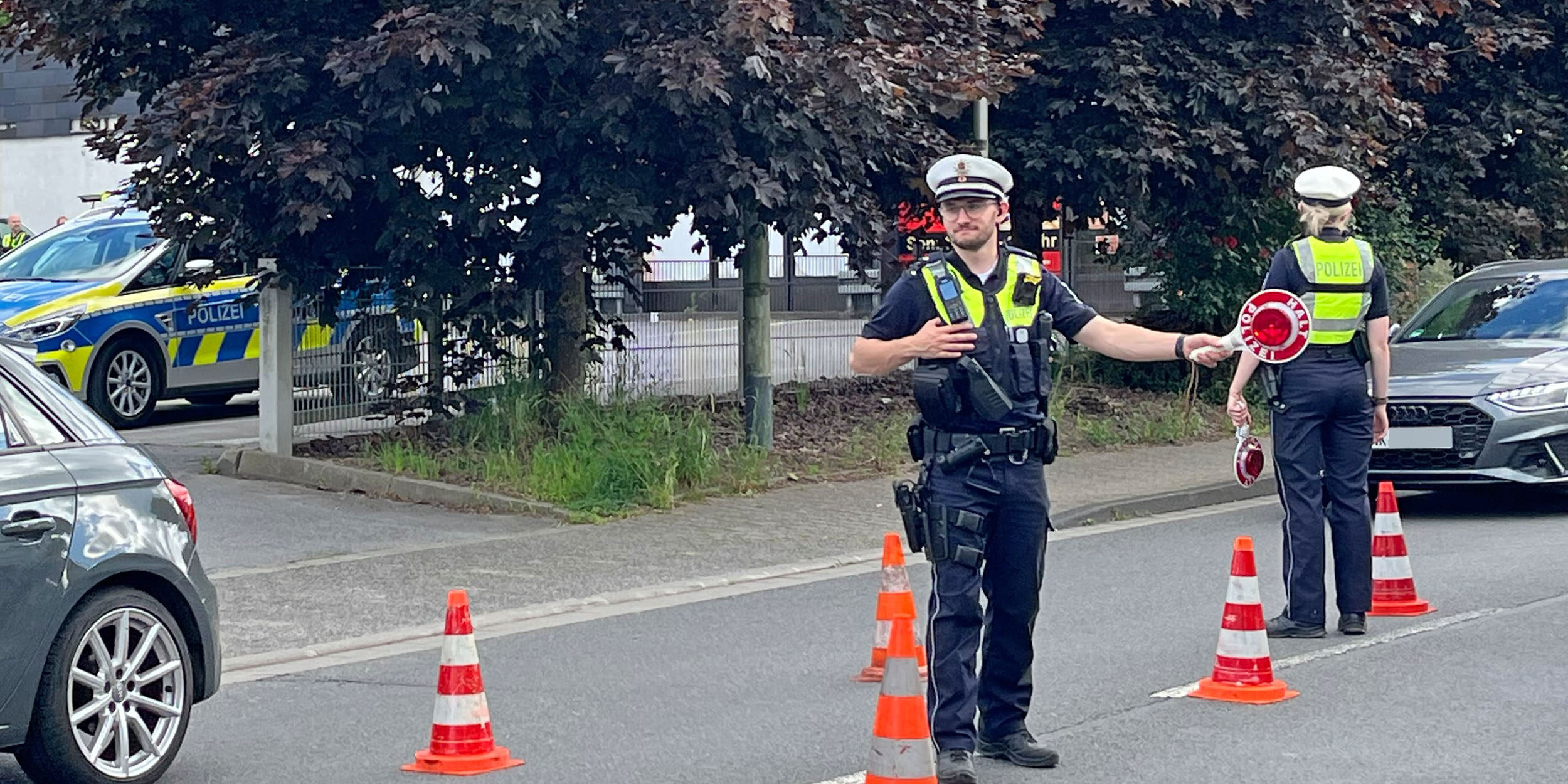 Zwei Polizeibeamte führen auf einer Straße eine Verkehrskontrolle durch. Zwischen mehreren orange-weißen Leitkegeln regeln sie den Verkehr und weisen Fahrzeuge ein. Im Hintergrund sind ein Streifenwagen sowie weitere Autos zu sehen. Die Aufnahme zeigt eine polizeiliche Kontrollstelle im öffentlichen Straßenverkehr.