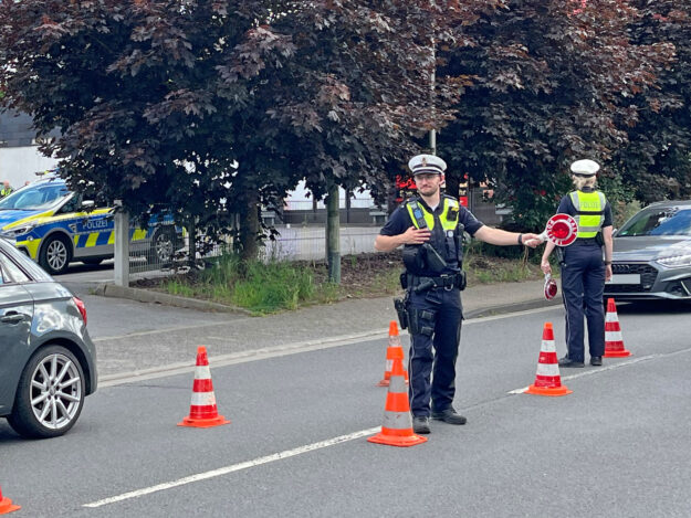 Zwei Polizeibeamte führen auf einer Straße eine Verkehrskontrolle durch. Zwischen mehreren orange-weißen Leitkegeln regeln sie den Verkehr und weisen Fahrzeuge ein. Im Hintergrund sind ein Streifenwagen sowie weitere Autos zu sehen. Die Aufnahme zeigt eine polizeiliche Kontrollstelle im öffentlichen Straßenverkehr.