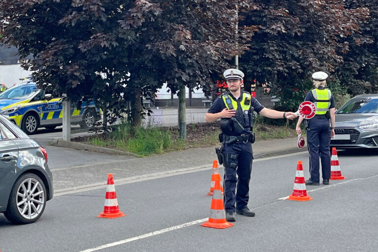 Zwei Polizeibeamte führen auf einer Straße eine Verkehrskontrolle durch. Zwischen mehreren orange-weißen Leitkegeln regeln sie den Verkehr und weisen Fahrzeuge ein. Im Hintergrund sind ein Streifenwagen sowie weitere Autos zu sehen. Die Aufnahme zeigt eine polizeiliche Kontrollstelle im öffentlichen Straßenverkehr.