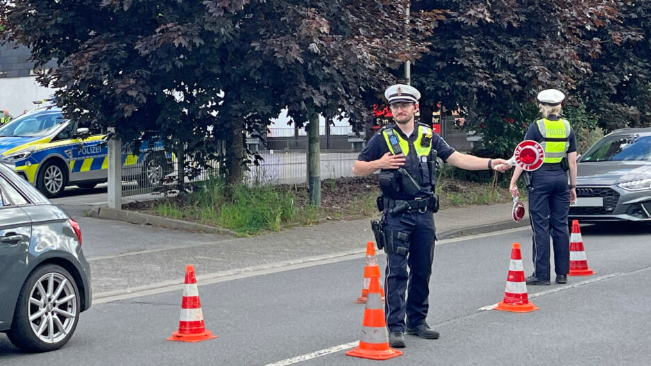 Zwei Polizeibeamte führen auf einer Straße eine Verkehrskontrolle durch. Zwischen mehreren orange-weißen Leitkegeln regeln sie den Verkehr und weisen Fahrzeuge ein. Im Hintergrund sind ein Streifenwagen sowie weitere Autos zu sehen. Die Aufnahme zeigt eine polizeiliche Kontrollstelle im öffentlichen Straßenverkehr.