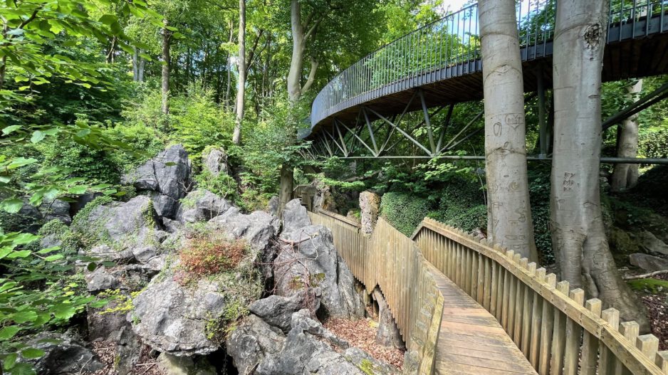 Ein hölzerner Steg führt durch eine felsige Waldlandschaft unterhalb einer geschwungenen Stahlbrücke. Hohe Bäume, große Felsformationen und dichtes Grün prägen die naturnahe Szenerie.