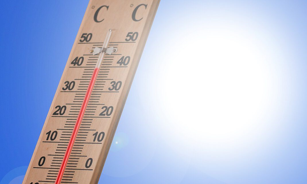 Ein Holzthermometer zeigt an einem sonnigen Tag eine Temperatur von rund 40 Grad Celsius an. Im Hintergrund strahlt eine helle Sonne vor einem wolkenlosen blauen Himmel. Die Aufnahme symbolisiert starke Sommerhitze und hohe Temperaturen.