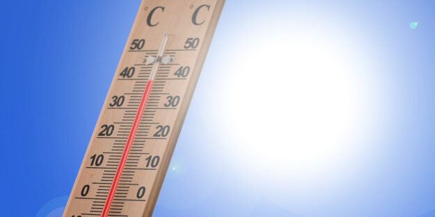 Ein Holzthermometer zeigt an einem sonnigen Tag eine Temperatur von rund 40 Grad Celsius an. Im Hintergrund strahlt eine helle Sonne vor einem wolkenlosen blauen Himmel. Die Aufnahme symbolisiert starke Sommerhitze und hohe Temperaturen.