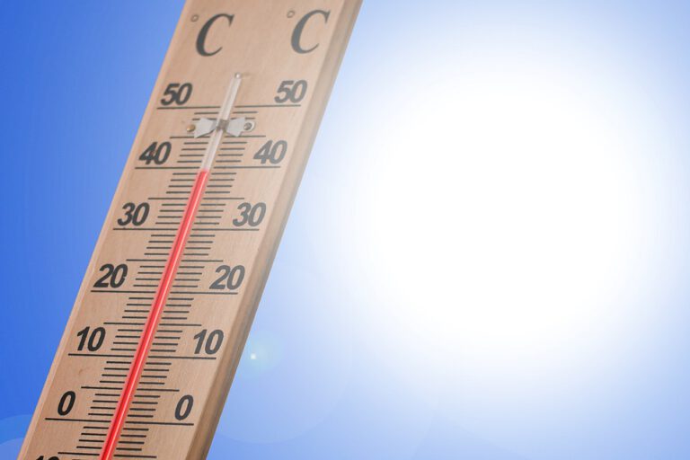 Ein Holzthermometer zeigt an einem sonnigen Tag eine Temperatur von rund 40 Grad Celsius an. Im Hintergrund strahlt eine helle Sonne vor einem wolkenlosen blauen Himmel. Die Aufnahme symbolisiert starke Sommerhitze und hohe Temperaturen.