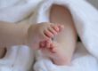 Nahaufnahme der kleinen Füße eines Babys, das in ein weiches weißes Handtuch eingewickelt ist. Die Zehen stehen im Mittelpunkt des Bildes, während der restliche Körper unscharf im Hintergrund zu sehen ist. Die Aufnahme vermittelt Geborgenheit und den Eindruck eines Neugeborenen oder Säuglings.