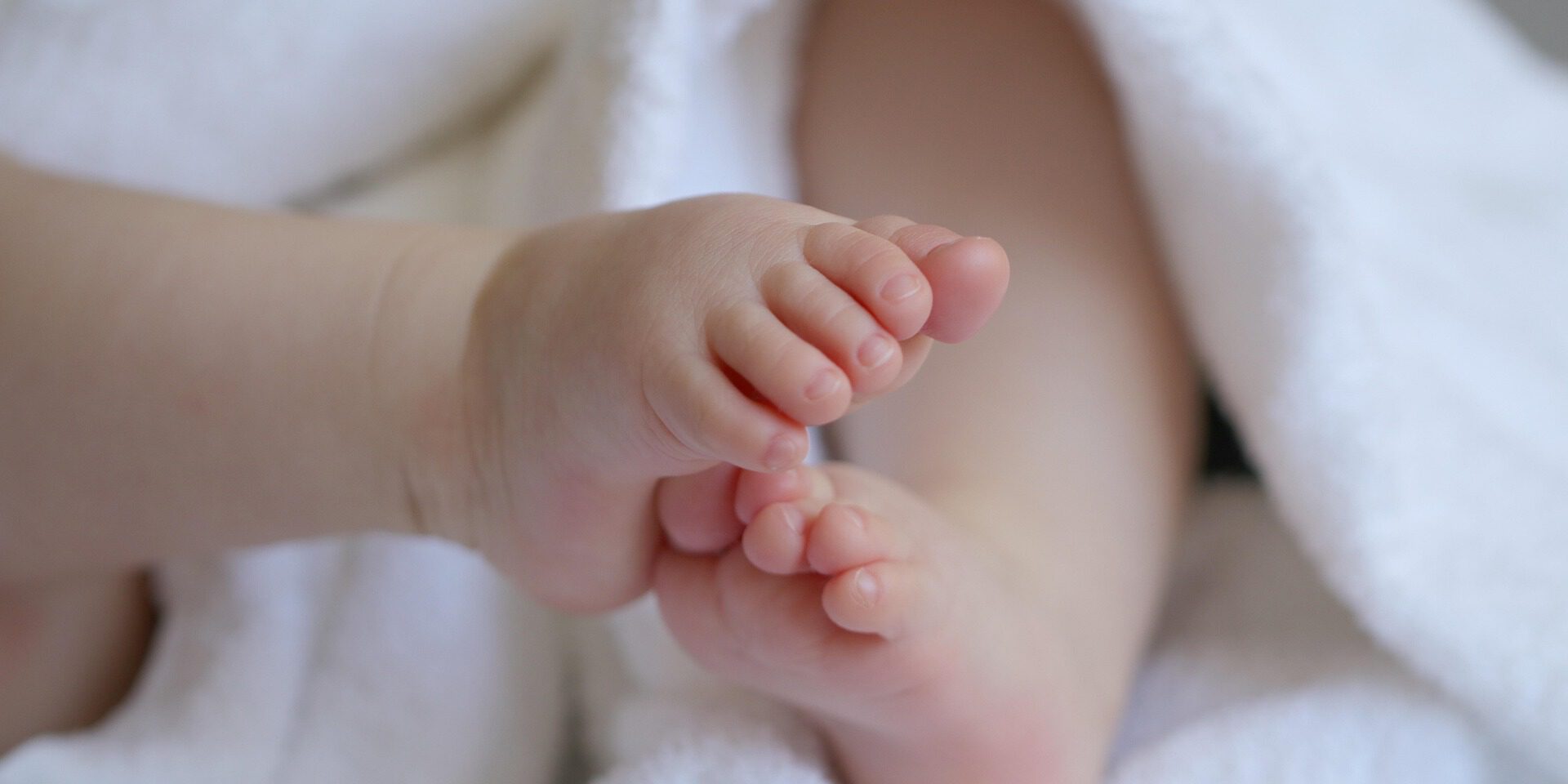 Nahaufnahme der kleinen Füße eines Babys, das in ein weiches weißes Handtuch eingewickelt ist. Die Zehen stehen im Mittelpunkt des Bildes, während der restliche Körper unscharf im Hintergrund zu sehen ist. Die Aufnahme vermittelt Geborgenheit und den Eindruck eines Neugeborenen oder Säuglings.