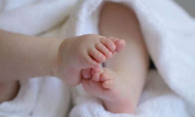 Nahaufnahme der kleinen Füße eines Babys, das in ein weiches weißes Handtuch eingewickelt ist. Die Zehen stehen im Mittelpunkt des Bildes, während der restliche Körper unscharf im Hintergrund zu sehen ist. Die Aufnahme vermittelt Geborgenheit und den Eindruck eines Neugeborenen oder Säuglings.
