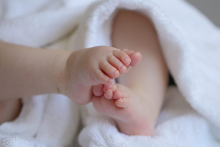 Nahaufnahme der kleinen Füße eines Babys, das in ein weiches weißes Handtuch eingewickelt ist. Die Zehen stehen im Mittelpunkt des Bildes, während der restliche Körper unscharf im Hintergrund zu sehen ist. Die Aufnahme vermittelt Geborgenheit und den Eindruck eines Neugeborenen oder Säuglings.