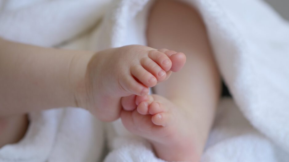Nahaufnahme der kleinen Füße eines Babys, das in ein weiches weißes Handtuch eingewickelt ist. Die Zehen stehen im Mittelpunkt des Bildes, während der restliche Körper unscharf im Hintergrund zu sehen ist. Die Aufnahme vermittelt Geborgenheit und den Eindruck eines Neugeborenen oder Säuglings.