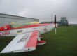 Ein rot-weiß lackiertes Kunstflugzeug steht auf einer Rasenfläche vor einem Hangar und dem Tower eines kleinen Flugplatzes. Der Propeller ruht, während der Himmel von tief hängenden grauen Wolken bedeckt ist. Die Aufnahme zeigt das Flugzeug aus seitlicher Perspektive und vermittelt die ruhige Atmosphäre eines Flugplatzes vor dem Start oder nach der Landung.