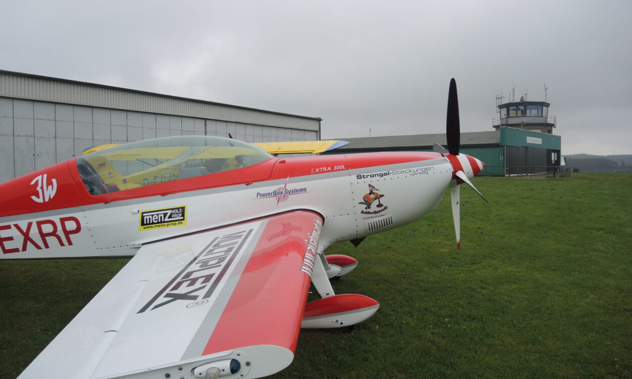 Ein rot-weiß lackiertes Kunstflugzeug steht auf einer Rasenfläche vor einem Hangar und dem Tower eines kleinen Flugplatzes. Der Propeller ruht, während der Himmel von tief hängenden grauen Wolken bedeckt ist. Die Aufnahme zeigt das Flugzeug aus seitlicher Perspektive und vermittelt die ruhige Atmosphäre eines Flugplatzes vor dem Start oder nach der Landung.