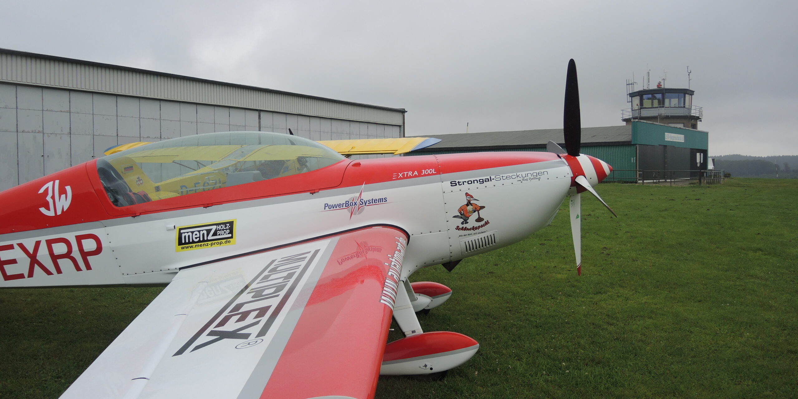 Ein rot-weiß lackiertes Kunstflugzeug steht auf einer Rasenfläche vor einem Hangar und dem Tower eines kleinen Flugplatzes. Der Propeller ruht, während der Himmel von tief hängenden grauen Wolken bedeckt ist. Die Aufnahme zeigt das Flugzeug aus seitlicher Perspektive und vermittelt die ruhige Atmosphäre eines Flugplatzes vor dem Start oder nach der Landung.