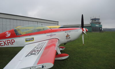 Ein rot-weiß lackiertes Kunstflugzeug steht auf einer Rasenfläche vor einem Hangar und dem Tower eines kleinen Flugplatzes. Der Propeller ruht, während der Himmel von tief hängenden grauen Wolken bedeckt ist. Die Aufnahme zeigt das Flugzeug aus seitlicher Perspektive und vermittelt die ruhige Atmosphäre eines Flugplatzes vor dem Start oder nach der Landung.