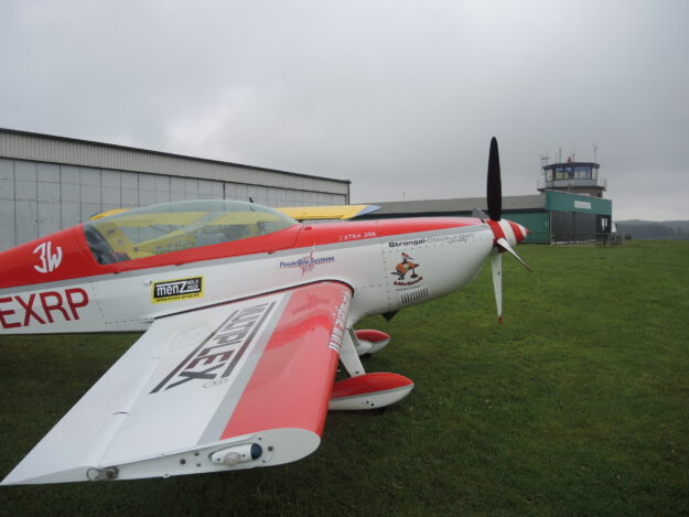 Ein rot-weiß lackiertes Kunstflugzeug steht auf einer Rasenfläche vor einem Hangar und dem Tower eines kleinen Flugplatzes. Der Propeller ruht, während der Himmel von tief hängenden grauen Wolken bedeckt ist. Die Aufnahme zeigt das Flugzeug aus seitlicher Perspektive und vermittelt die ruhige Atmosphäre eines Flugplatzes vor dem Start oder nach der Landung.