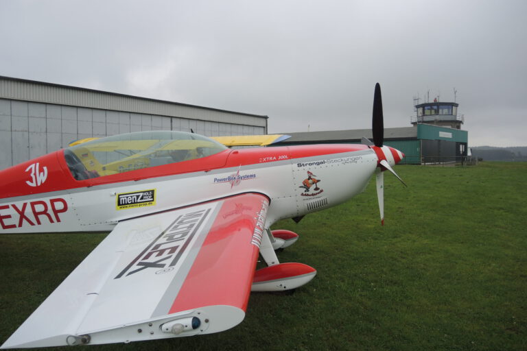 Ein rot-weiß lackiertes Kunstflugzeug steht auf einer Rasenfläche vor einem Hangar und dem Tower eines kleinen Flugplatzes. Der Propeller ruht, während der Himmel von tief hängenden grauen Wolken bedeckt ist. Die Aufnahme zeigt das Flugzeug aus seitlicher Perspektive und vermittelt die ruhige Atmosphäre eines Flugplatzes vor dem Start oder nach der Landung.